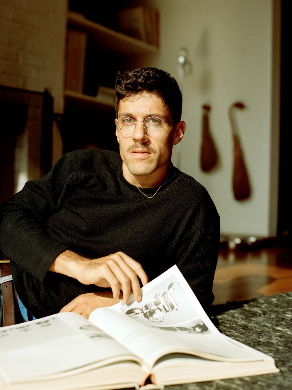 Adult male with glasses sitting indoors looking at an open book on a table, wearing a black sweater, in a naturally lit setting.