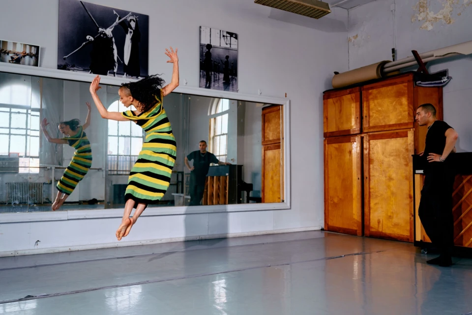 Female dancer leaps in studio, reflected in mirror with male observer; wooden cabinets, piano, and artwork enhance the backdrop.