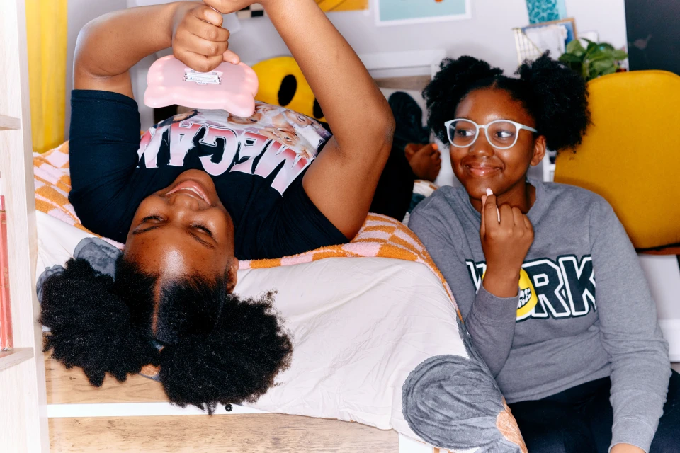 Two happy African American teenage girls pose indoors in a bedroom setting, one upside down on the bed, smiling with a toy.