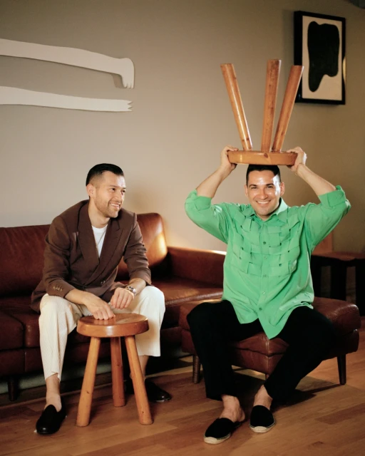 Two adult males pose indoors on a brown leather couch. One man balances a wooden stool on his head while smiling.
