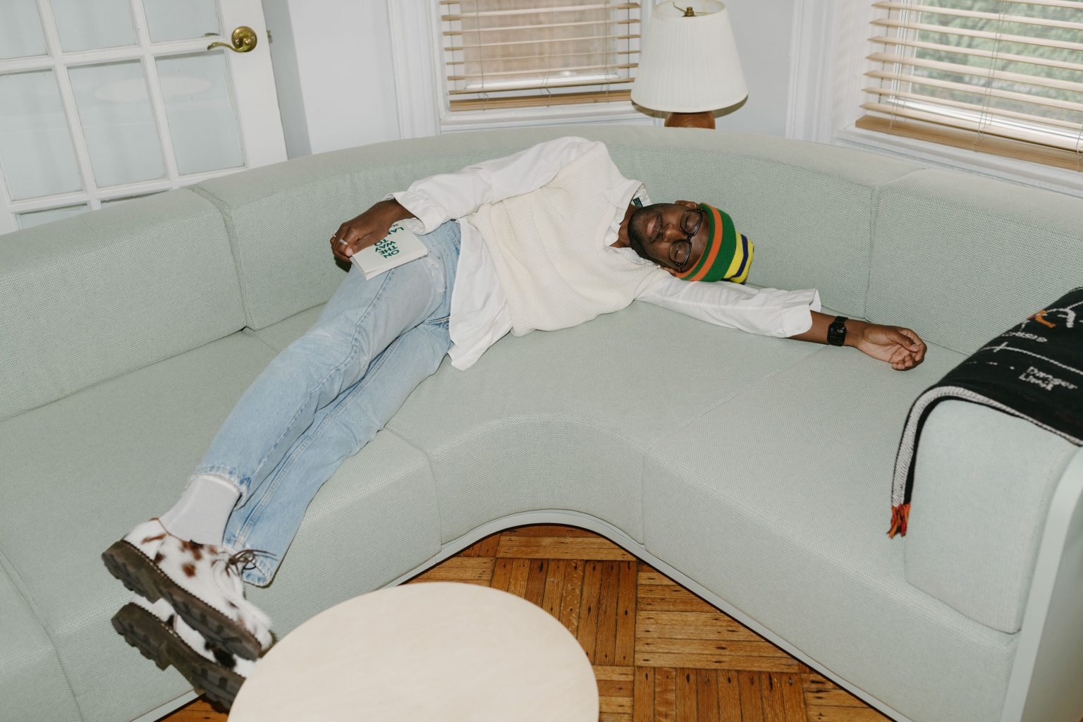 Man lounges on a light blue couch in casual outfit, wearing a colorful hat, holding a book in a brightly lit indoor setting.
