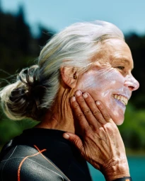 Profile portrait of an elderly woman with silver hair applying sunscreen to her cheek outdoors, captured by Caroline Tompkins