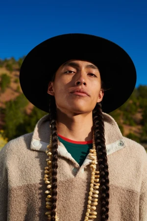 Portrait of a man with long braided dark hair wearing a black wide-brimmed hat, beige fleece pullover, and shell necklace against a clear blue sky