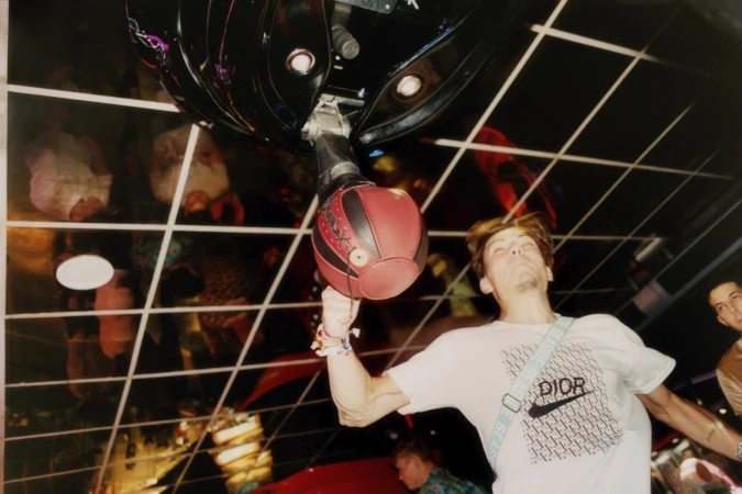 Adult male playing with red basketball in an indoor venue with grid patterned ceiling and crowd. He is wearing a white Dior branded tee.
