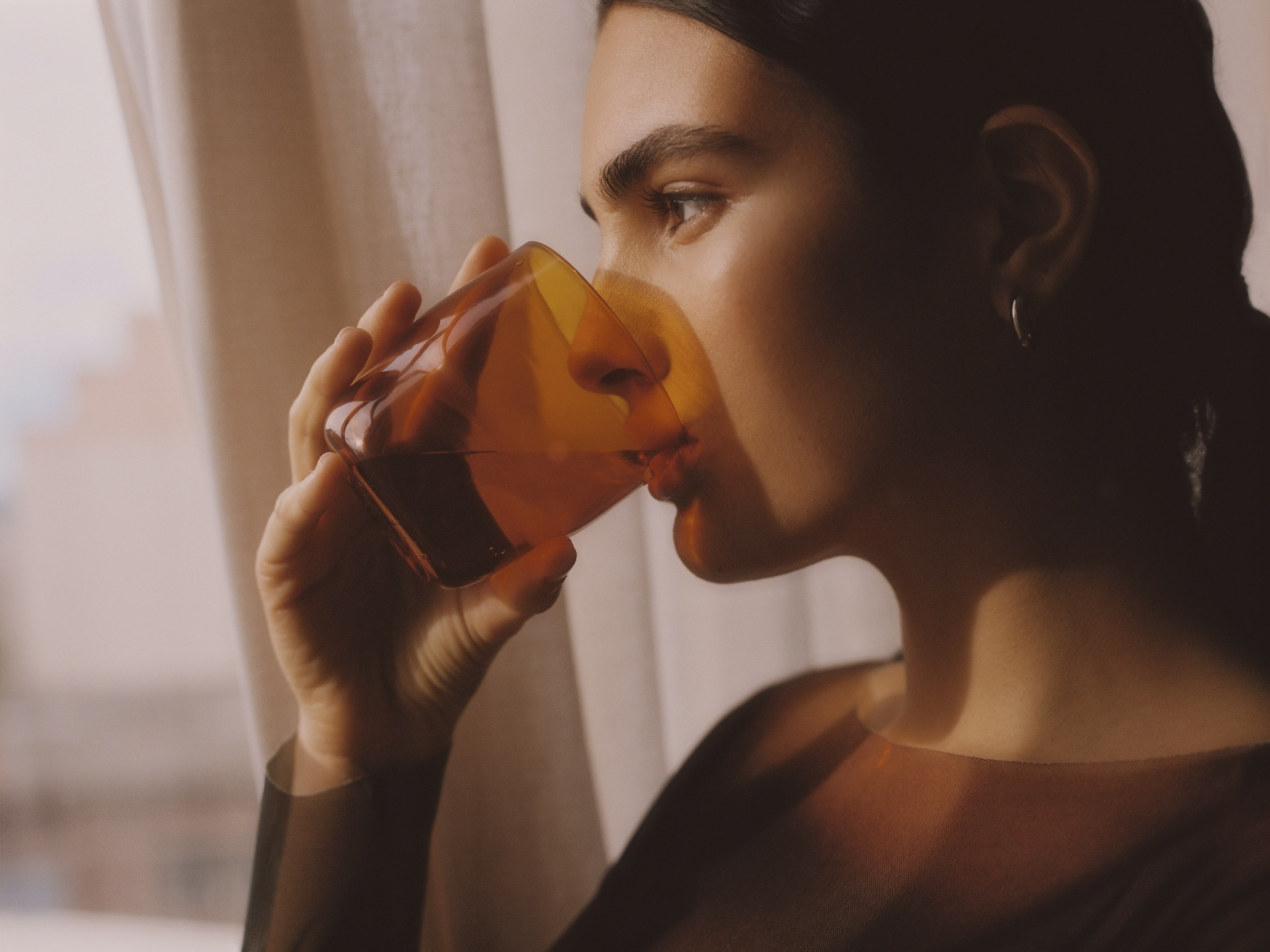 Close-up of a brunette woman with fair skin drinking from a textured amber glass in a soft, natural light setting near a window.