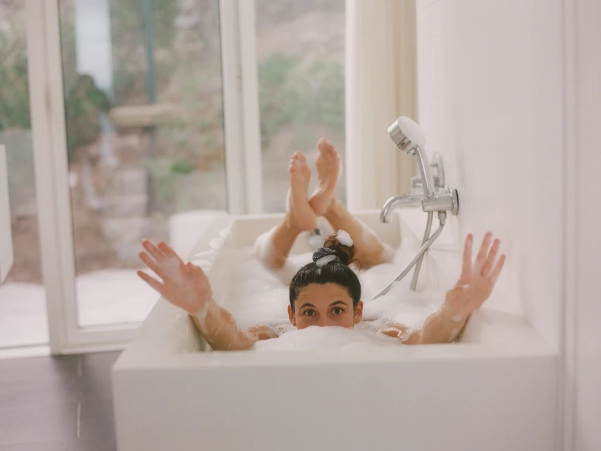 Woman relaxing in a bubble bath, raising her arms with a playful expression, light indoor setting with large window and outdoor view.