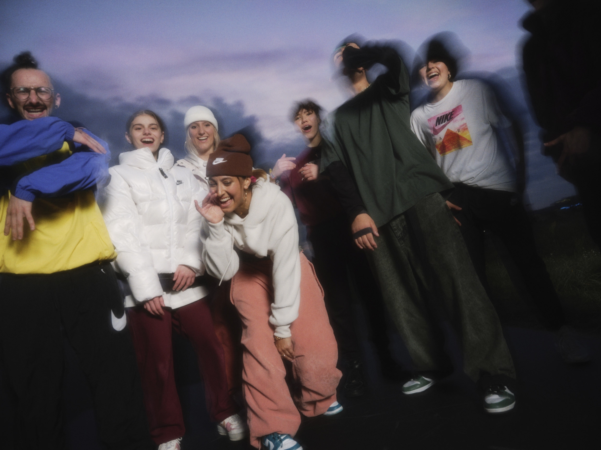 A group of seven people, including adult men and women, are smiling and posing together outdoors at dusk wearing casual clothing with a sky background.