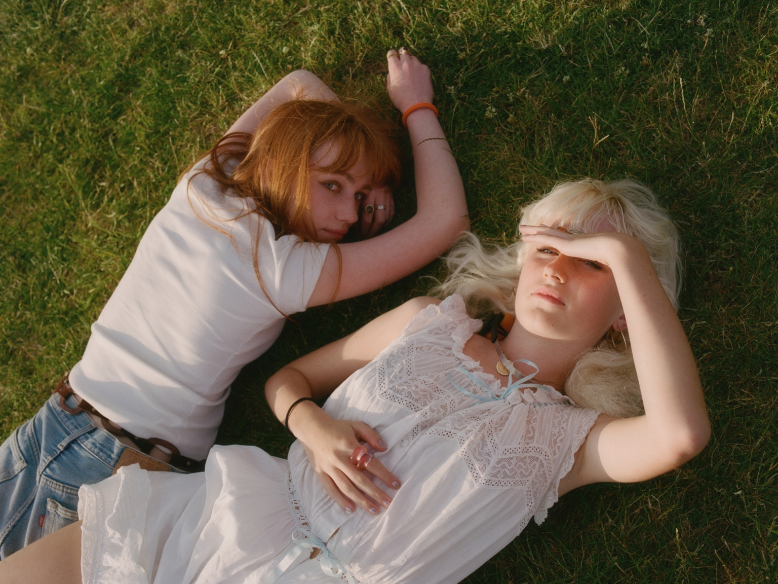 Two young women with red and blonde hair lying on green grass outdoors in a high angle portrait shot by Ana Cuba.