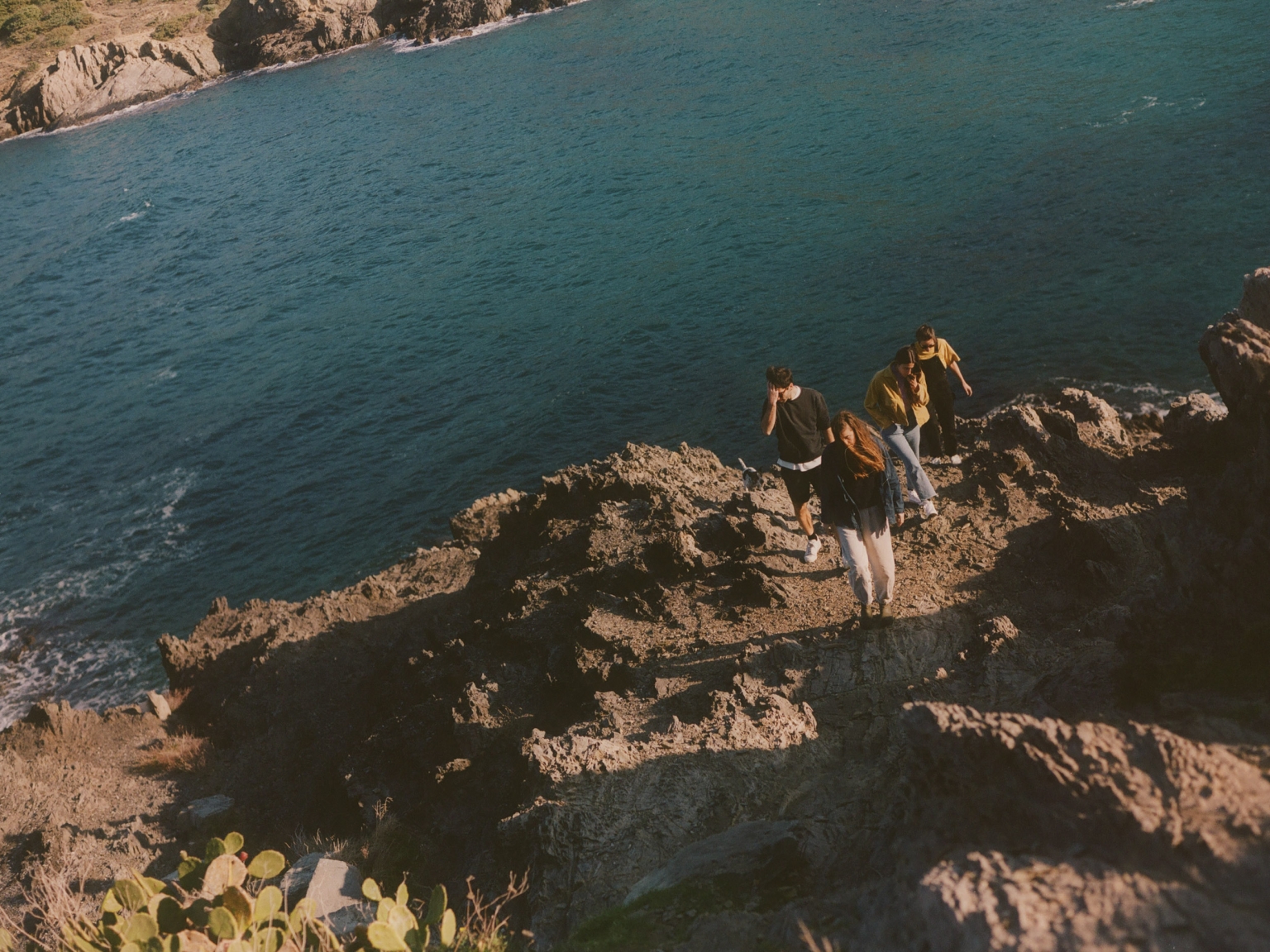 A group of four people standing on a rocky cliff overlooking a calm blue sea on a sunny day. The landscape is rugged and natural.
