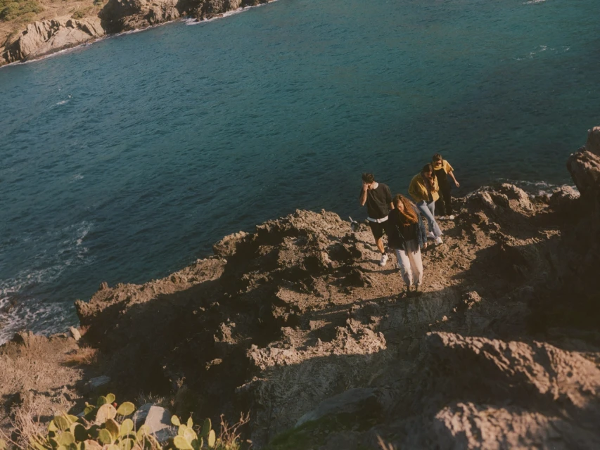 A group of four people standing on a rocky cliff overlooking a calm blue sea on a sunny day. The landscape is rugged and natural.