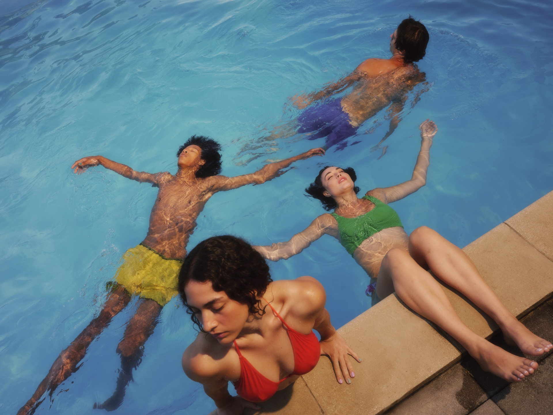 Four young adults relax in a light-blue swimming pool, wearing colorful swimwear, with one woman sitting on the edge of the pool.