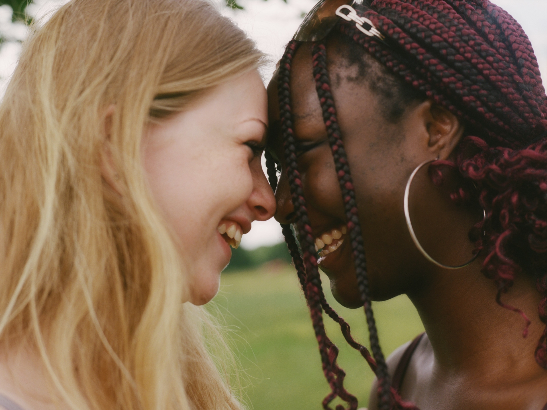 Two women, a blonde and a woman with dark braids, face each other smiling in an outdoor setting, heads almost touching, close-up shot.