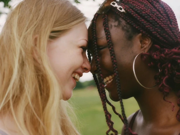 Two smiling women pressing their foreheads together outdoors against a soft green park background