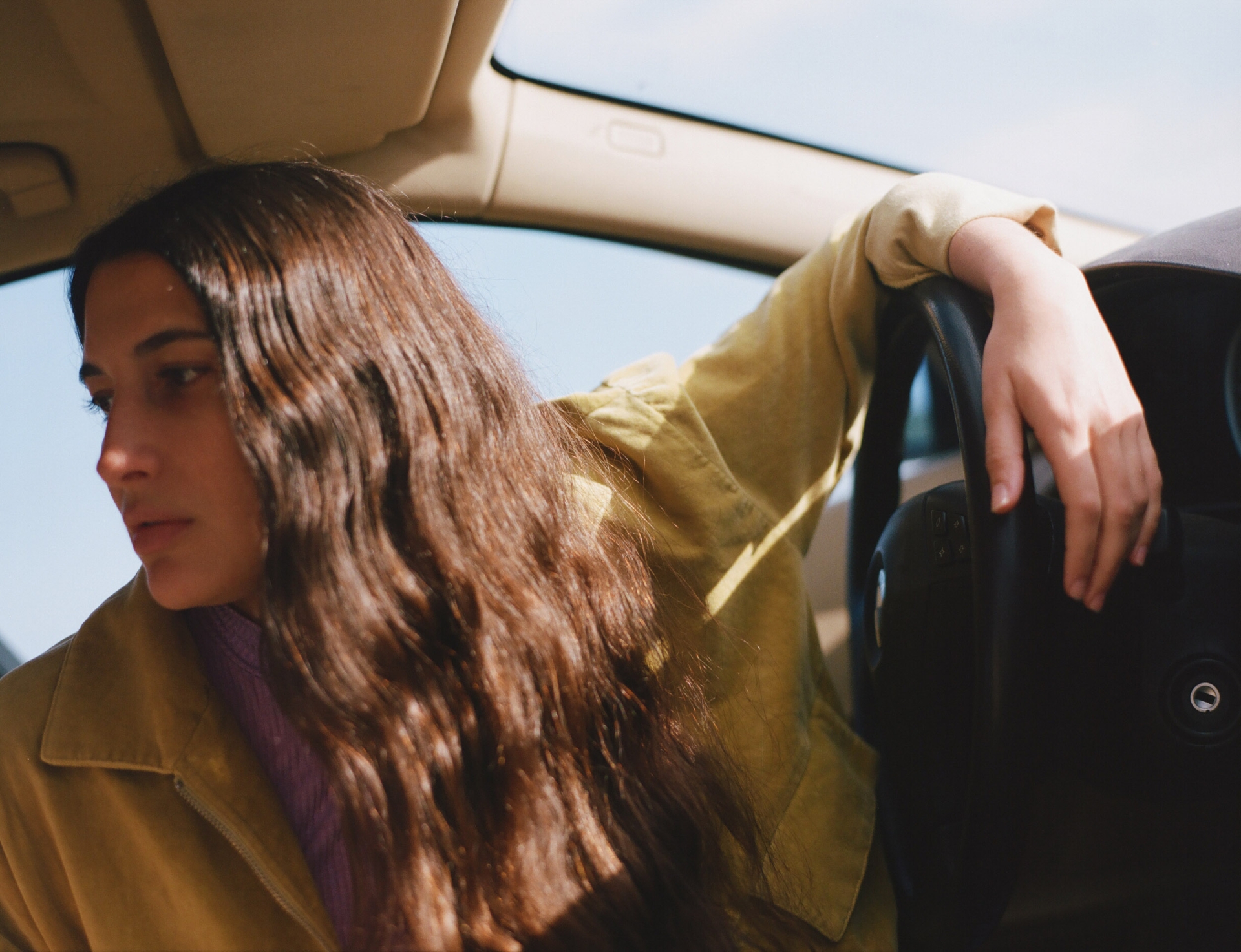 Woman with long brown hair relaxes in the driver's seat of a car on a sunny day, wearing a yellow jacket, gazing out the window.