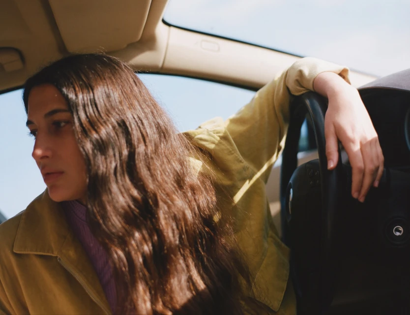 Woman with long brown hair relaxes in the driver's seat of a car on a sunny day, wearing a yellow jacket, gazing out the window.