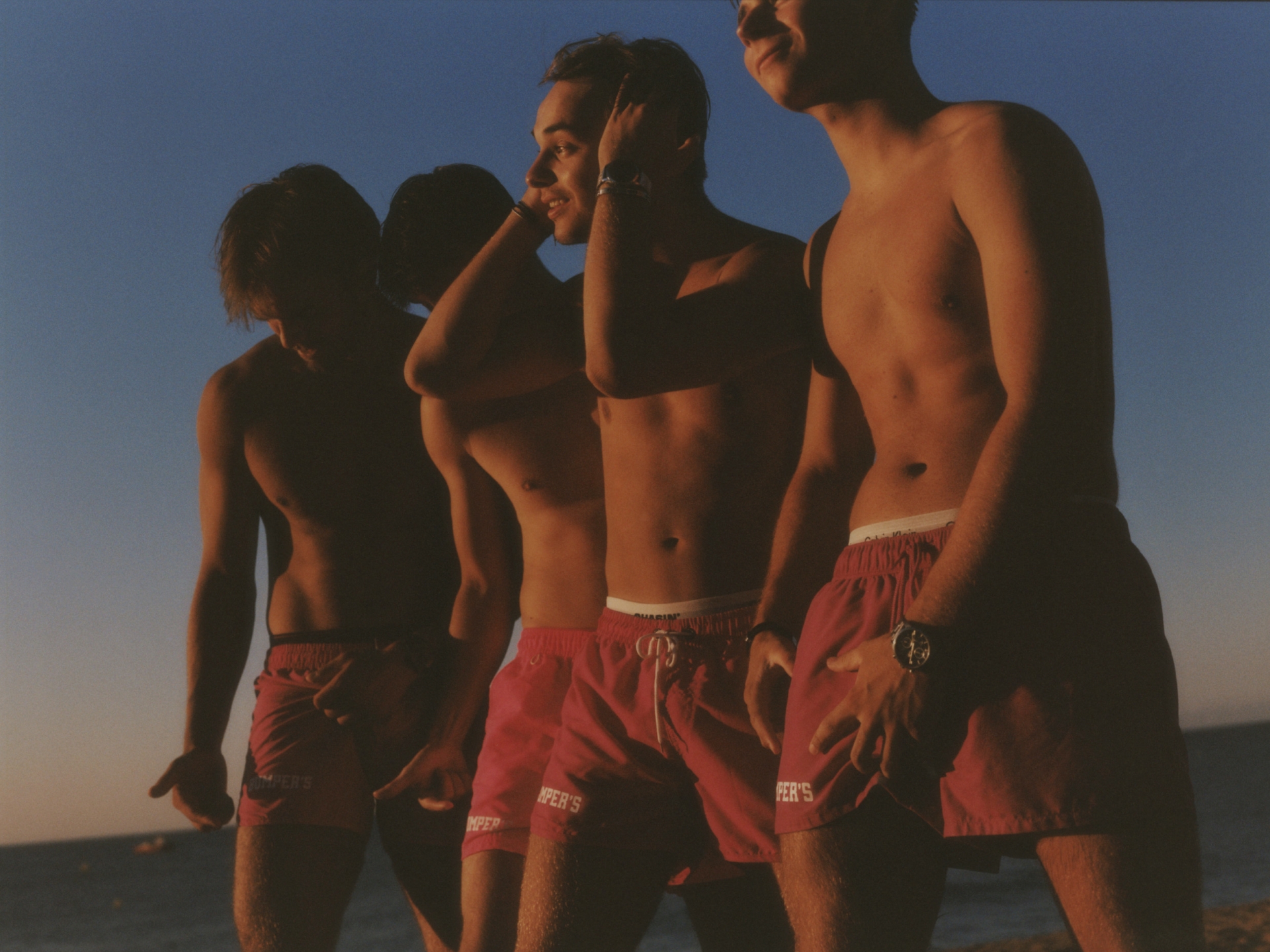 Four topless men in red swim trunks stand close together on a beach under a blue sky during sunset. Their bodies are tan.