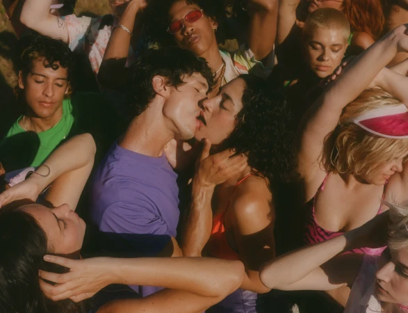 A group of diverse young adults gather outdoors, with a couple kissing in the foreground, surrounded by friends in a natural setting.