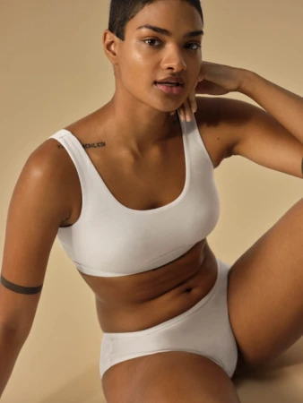 Brunette woman in white underwear sitting against beige background. Close-up of fair-skinned female in underwear, simple studio shot.