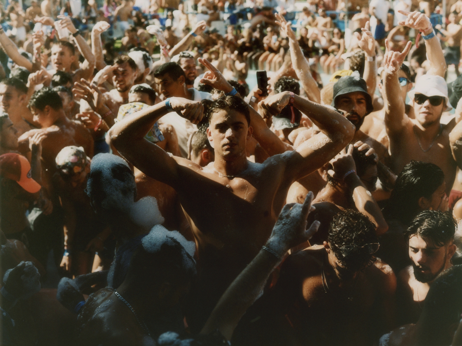Large crowd of shirtless young adults and teenagers gather outdoors in daylight, many with arms raised, appearing to cheer and celebrate.