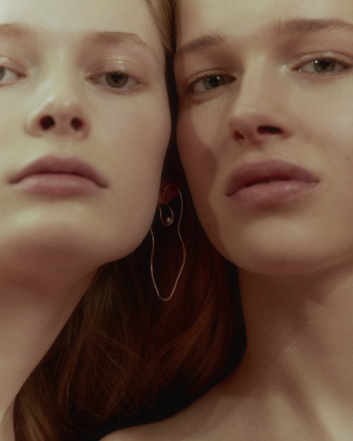 Close-up portrait of two young adult women with fair skin and light hair, one woman wearing a silver drop earring, in soft studio light.