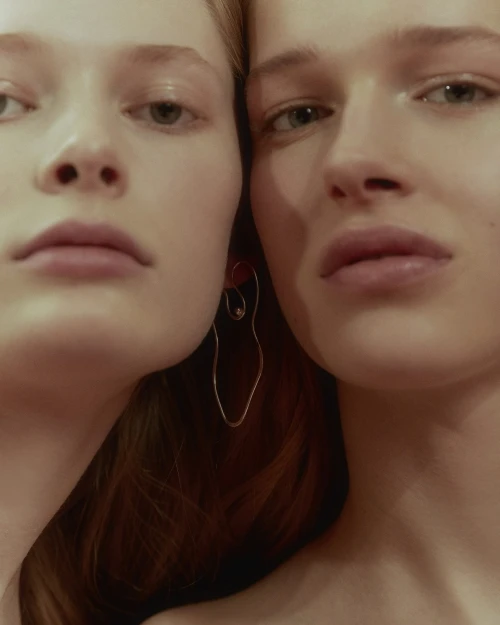 Close-up portrait of two young adult women with fair skin and light hair, one woman wearing a silver drop earring, in soft studio light.
