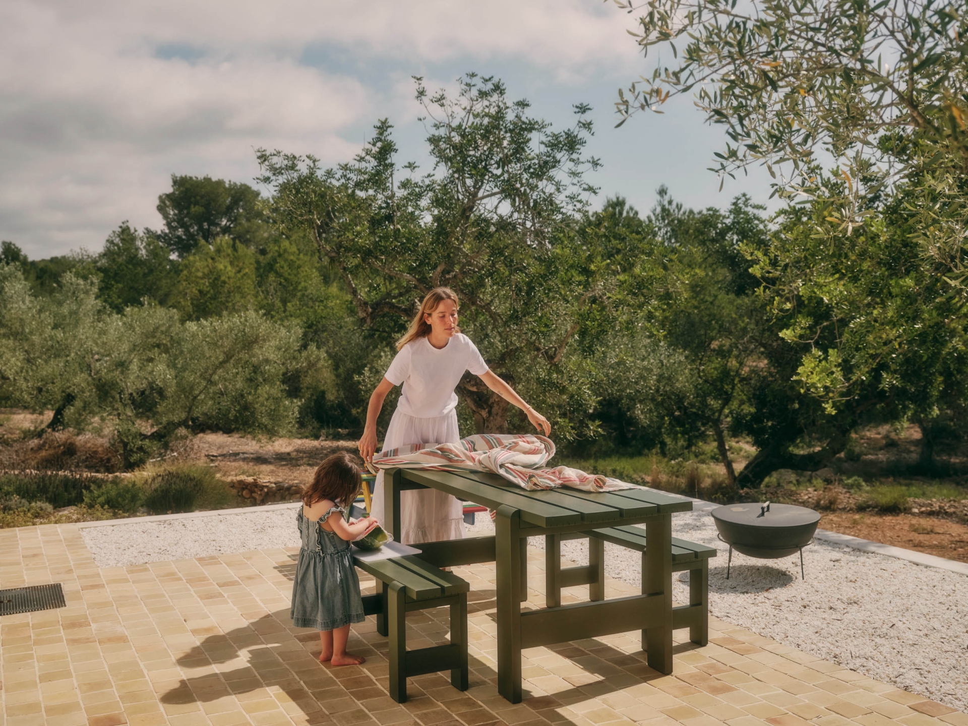 Woman and child at an olive-green picnic table on a stone patio with trees in the background on a sunny day, natural light.