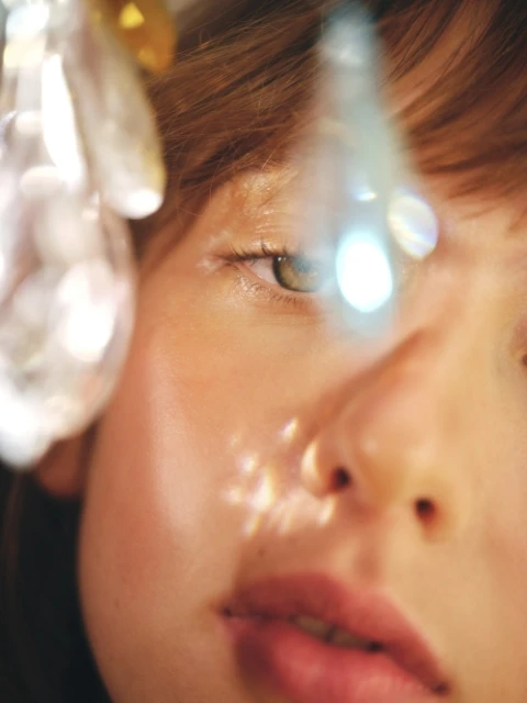 Close-up portrait of a woman's face with fair skin, light brown hair, and green eyes, featuring a shallow depth of field and crystal-like objects.