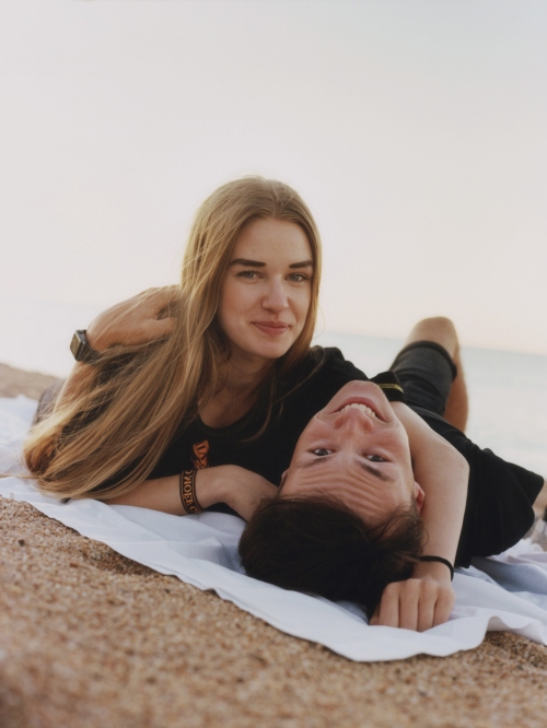 A young couple lounges together on a beach blanket against a soft, neutral background in an eye-level shot, creating a tranquil scene.
