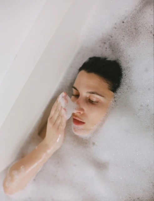 Brunette woman lies in a white bathtub filled with bubbles, with her eyes closed and a hand near her face in an indoor setting.