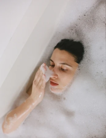 Brunette woman lies in a white bathtub filled with bubbles, with her eyes closed and a hand near her face in an indoor setting.