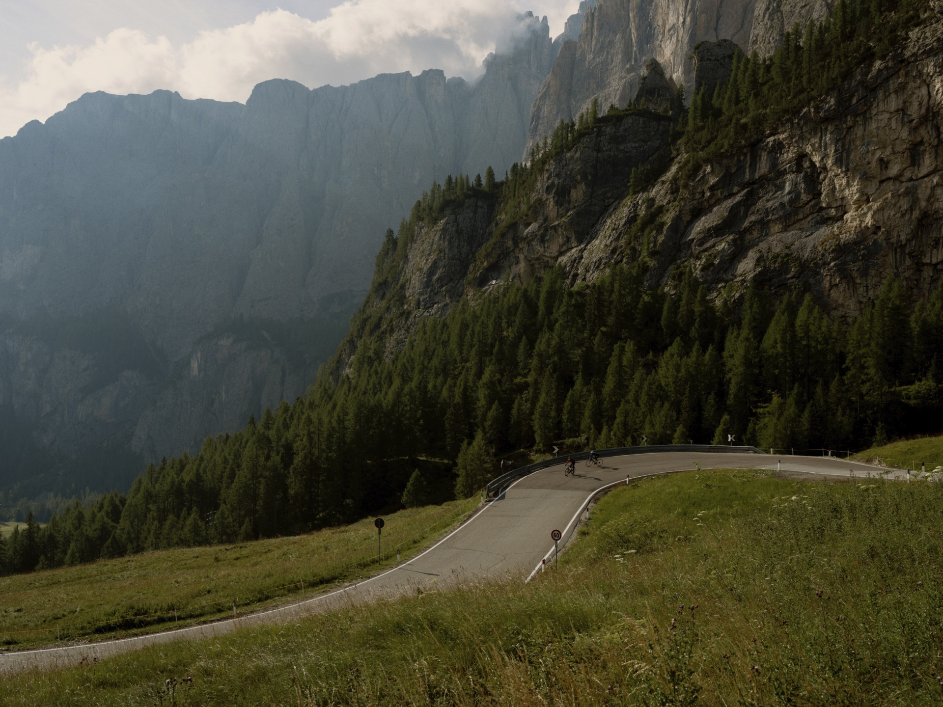 Scenic mountain road with cyclists winding through green grass and forest, set against a backdrop of rugged cliffs and cloudy sky.