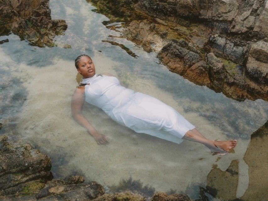 An adult female lies in shallow water amongst rocks in a light dress, basking in sunlight. The clear water adds a serene quality.