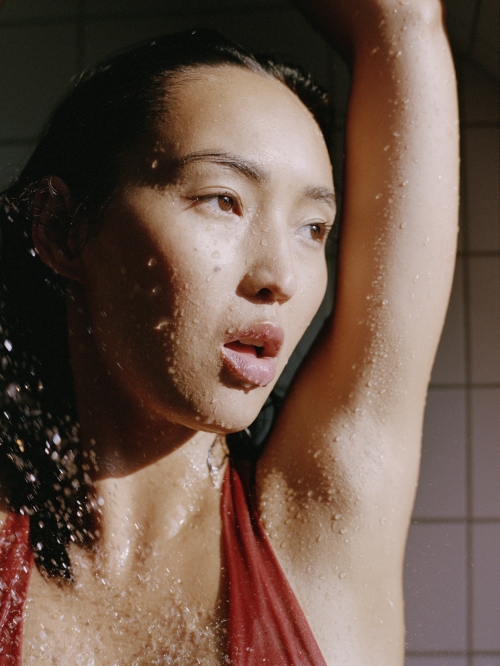 Close-up of a woman with an open mouth, posing in a wet swimsuit, with tiled wall visible in the background and light shining.