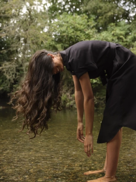 Woman in a black dress standing in a shallow river with trees and sky visible. Long brown hair hangs down toward the water.