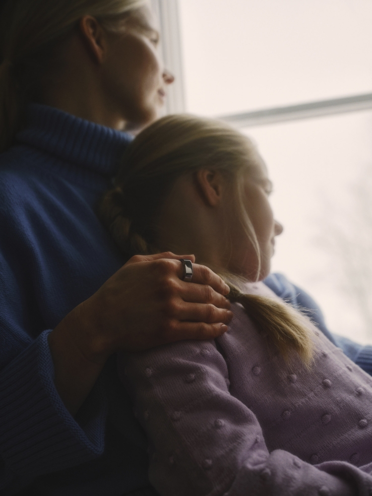 A mother and child embrace by a window; the mother is wearing a blue sweater and the child is in lilac; looking out the window.