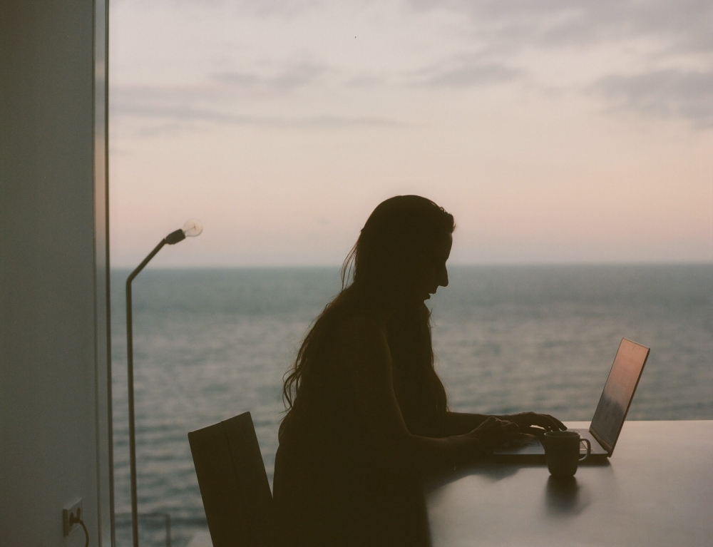 Silhouette of a woman working on a laptop at a table by a large window overlooking the ocean at dusk.