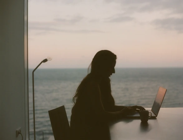 Silhouette of a woman working on a laptop at a table by a large window overlooking the ocean at dusk.
