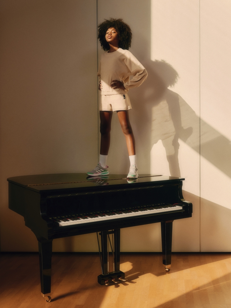 Adult female in streetwear poses confidently indoors with her foot on a piano, lit by warm, natural light in a fashion editorial.