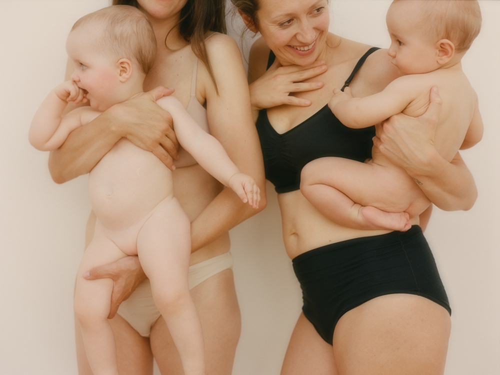 Two young women in underwear holding babies in a photo studio, smiling. The soft lighting creates a nurturing and familial atmosphere.