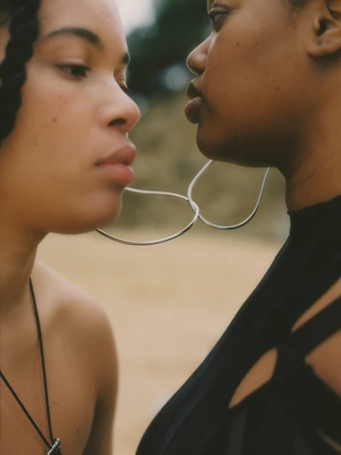 Close-up shot of two dark-skinned adult females with braided hair and black tops against a sandy background, showcasing a natural aesthetic.