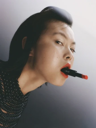 An Asian woman with red lips is holding a Lelo lipstick against her mouth in a studio setting with diffused lighting.