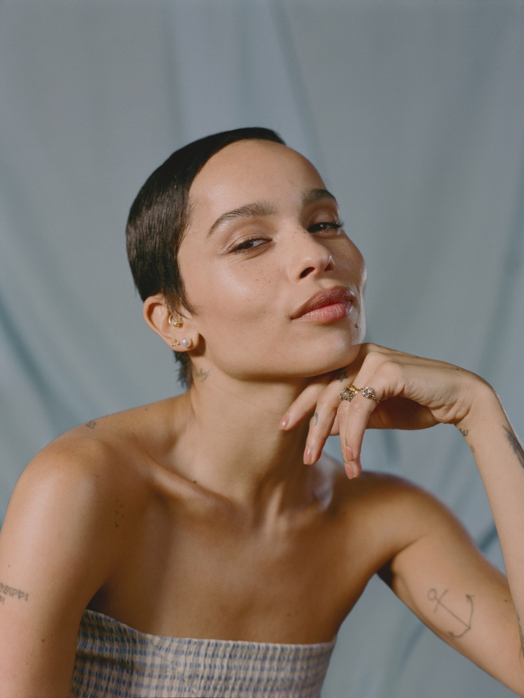 Portrait of Zoë Kravitz against a soft blue background. Kravitz has short hair, light skin, and is wearing a strapless checkered top.