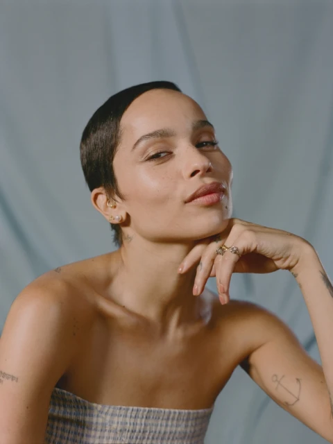 Portrait of Zoë Kravitz against a soft blue background. Kravitz has short hair, light skin, and is wearing a strapless checkered top.