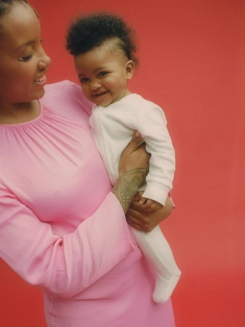 Mother holding a baby in a white onesie against a red background. The woman is wearing a long-sleeved pink dress with a gathered neckline.