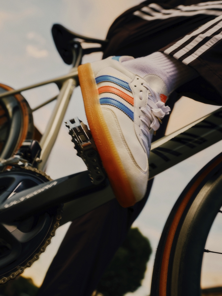Closeup of a person riding a bicycle, foot on pedal wearing a white Adidas Samba sneaker with blue and orange stripes.