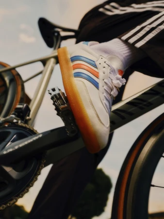 Closeup of a person riding a bicycle, foot on pedal wearing a white Adidas Samba sneaker with blue and orange stripes.