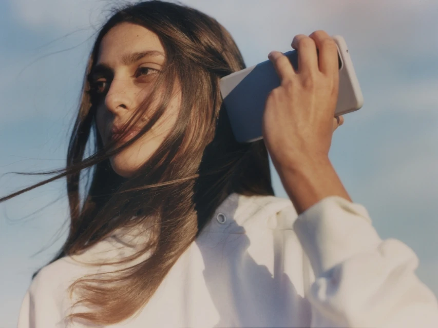 Portrait of a young woman with long hair holding a cell phone against a blurred blue sky, wearing a casual white top outdoors.