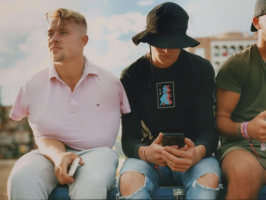 Three adult males sit outside on a sunny day; one uses a phone, and all wear casual clothing. Blue sky and a building are visible.