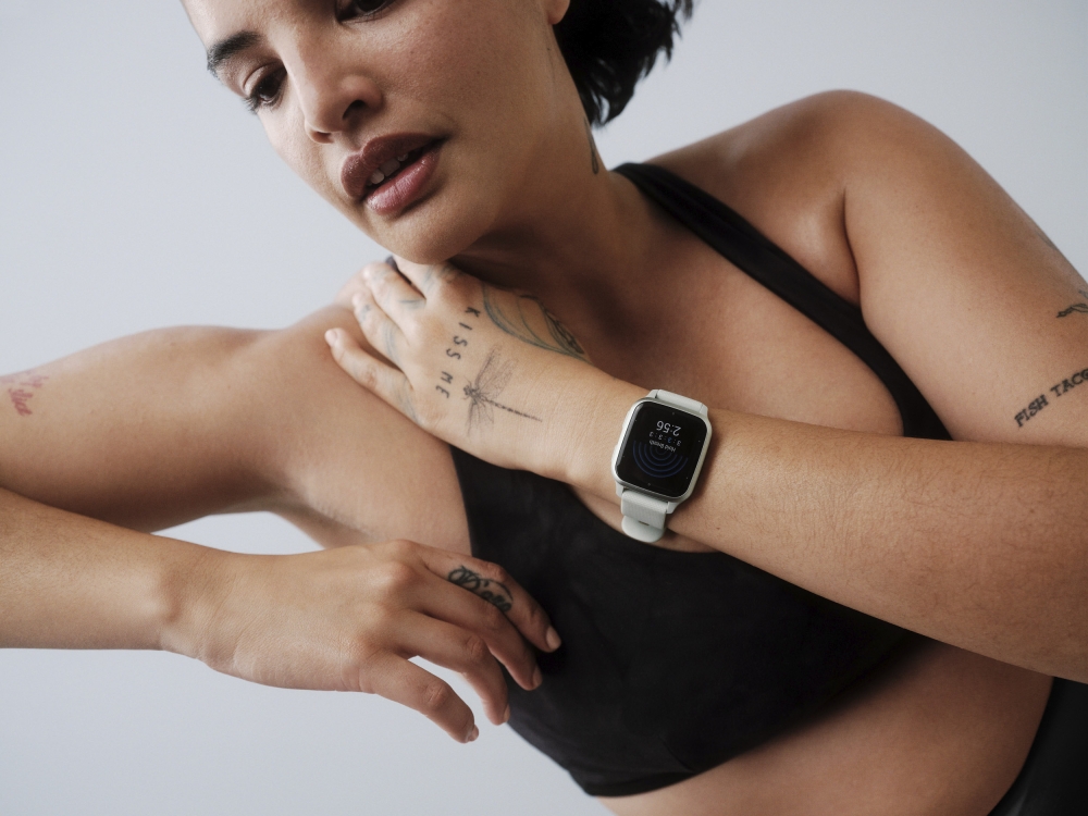 Woman wearing a black sports bra and a smart watch with white band, resting her hand on her shoulder in a bright, neutral setting.