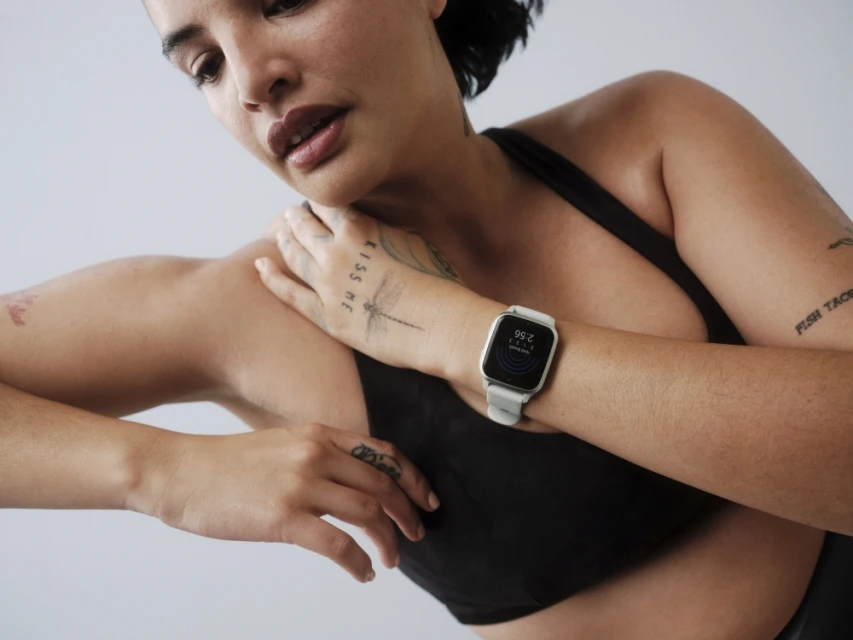 Woman wearing a black sports bra and a smart watch with white band, resting her hand on her shoulder in a bright, neutral setting.